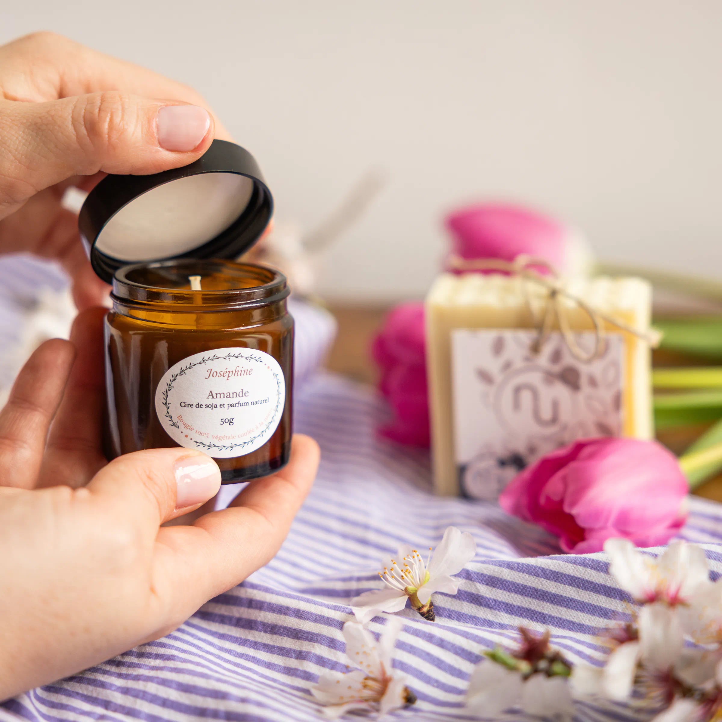 Hands opening Joséphine almond scented candle jar with Provençal soap in background