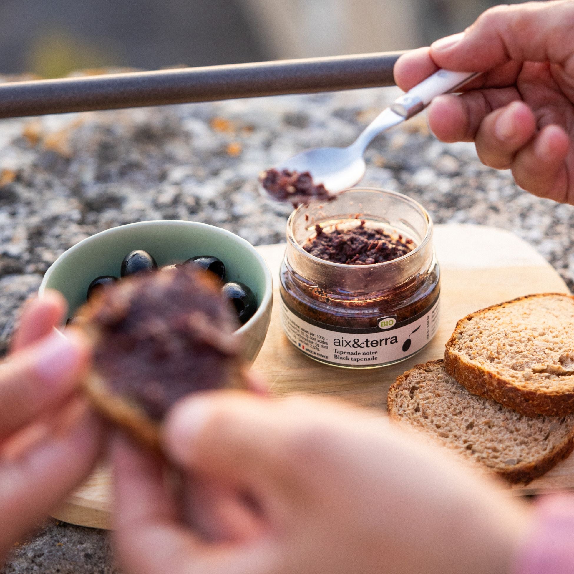 An authentic Provençal aperitivo ritual setup on a rustic wooden board, featuring a bottle of Céline en Provence extra virgin olive oil for dipping bread, a jar of organic black tapenade, and a bowl of olives and almonds, under warm, golden afternoon light.
