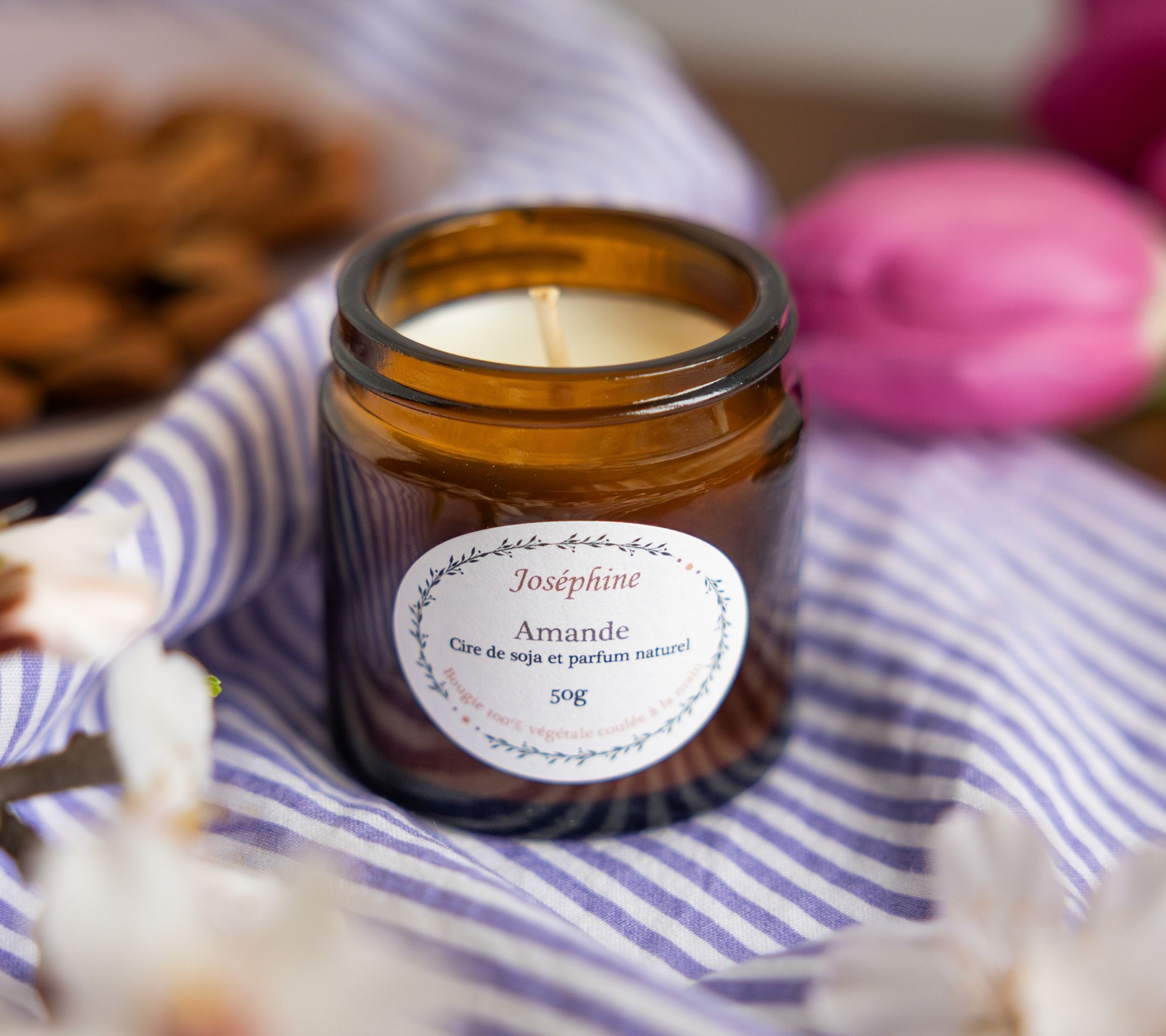 An amber glass jar of handcrafted almond scented candle resting on a blue and white striped fabric, surrounded by white spring blossoms, for a serene home ritual.
