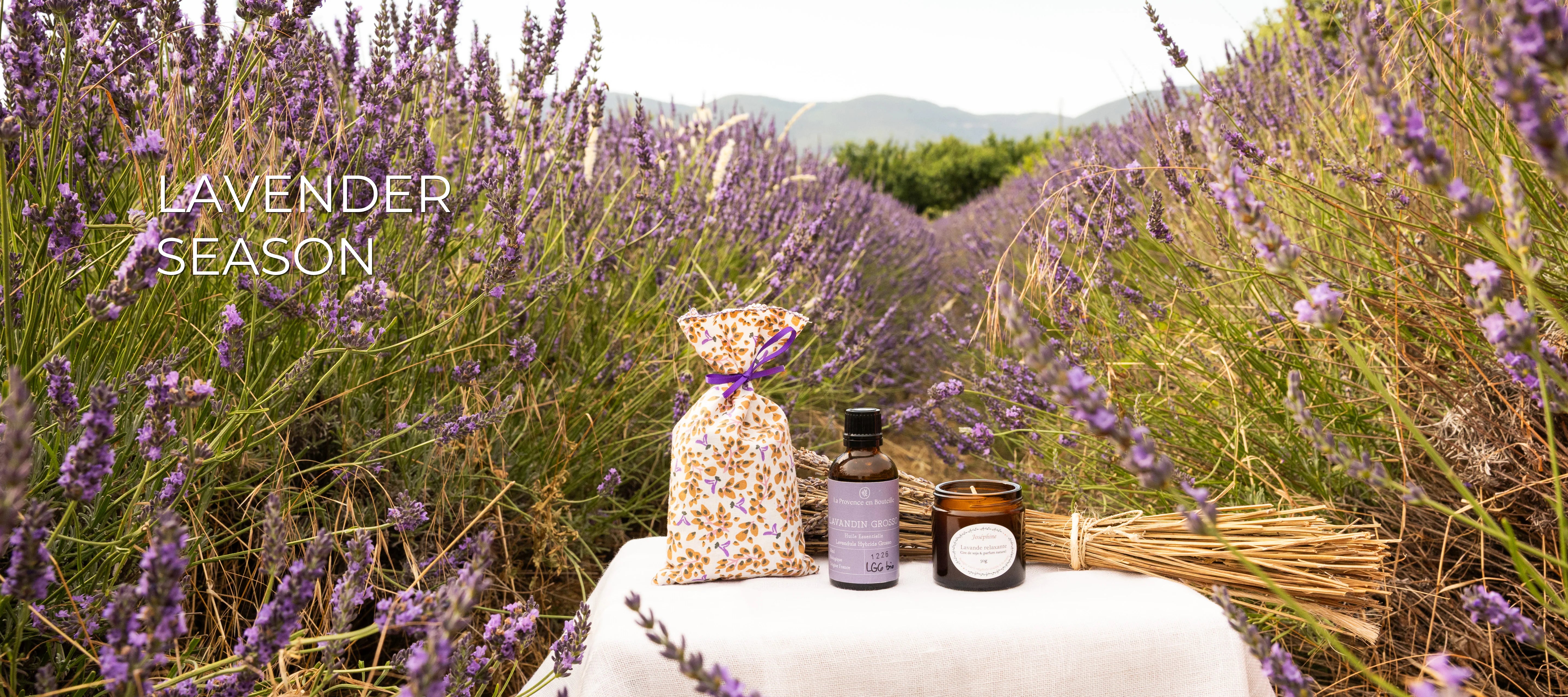 Lavender Season banner with lavender pouch, Lavandin Grosso essential oil and lavender scented candle on a linen table in a Provençal lavender field