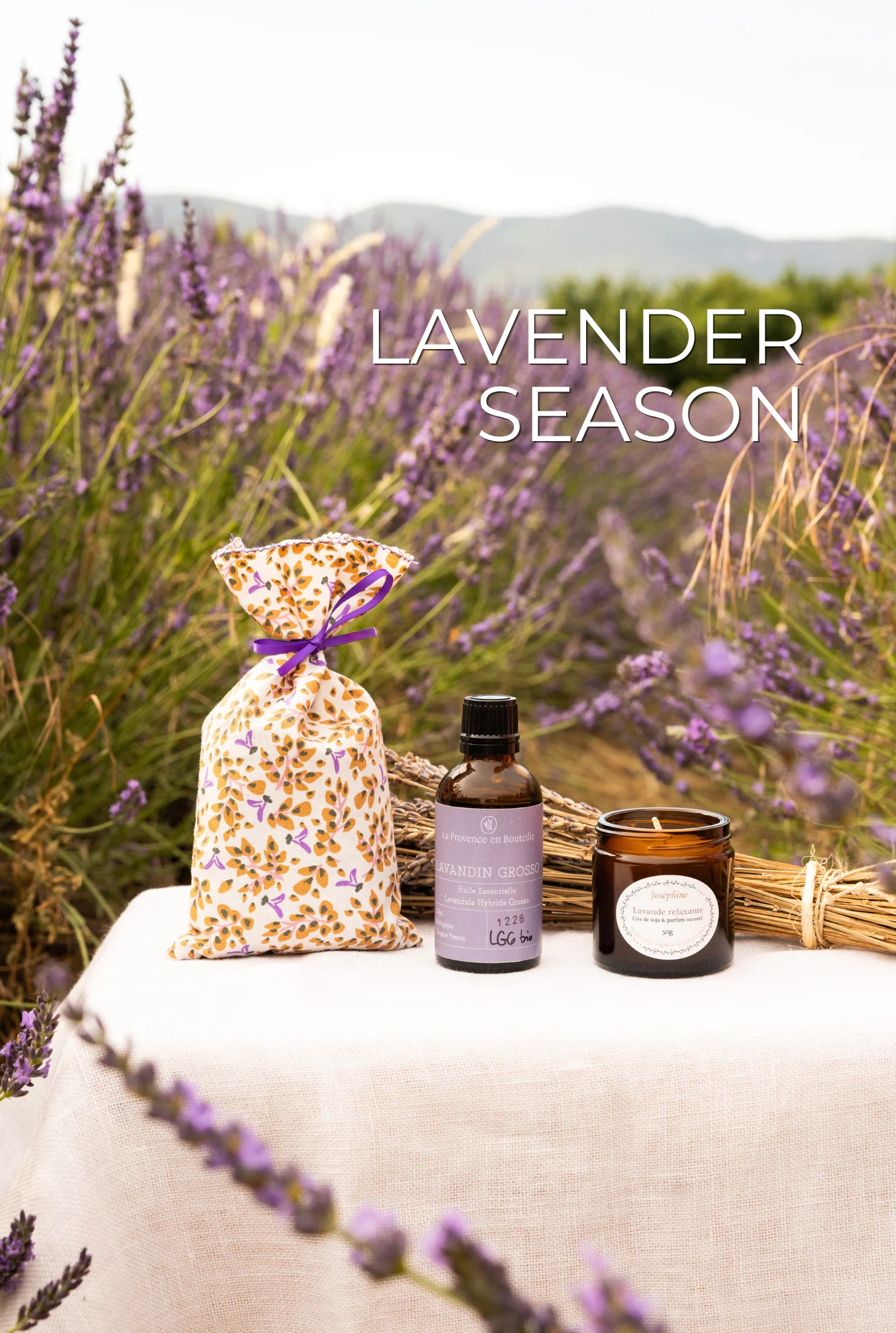 Lavender Season banner with lavender pouch, Lavandin Grosso essential oil and lavender scented candle on a linen table in a Provençal lavender field