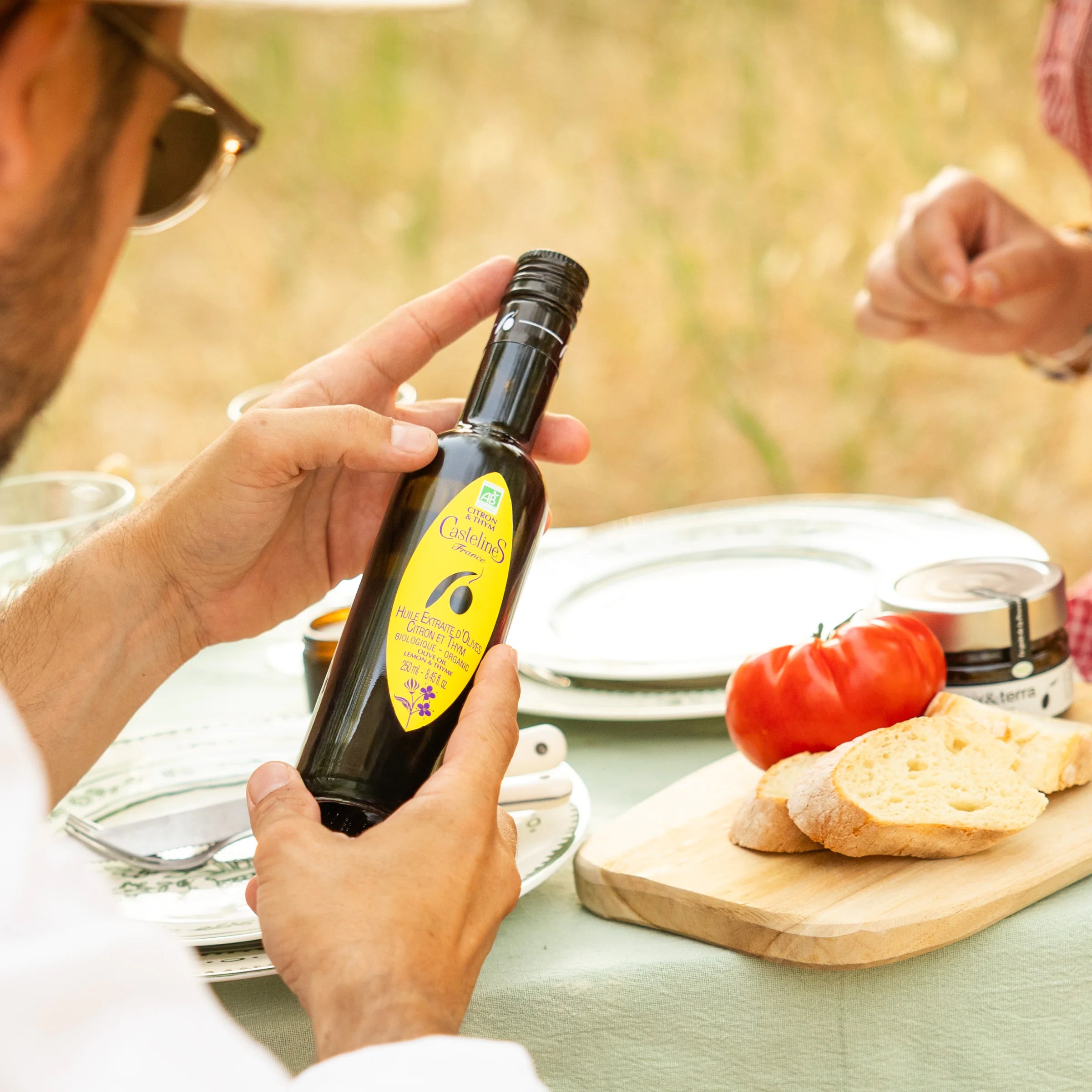 Male hands holding Castelines lemon olive oil bottle at an outdoor summer meal with tomatoes, bread and tapenade, Provençal table
