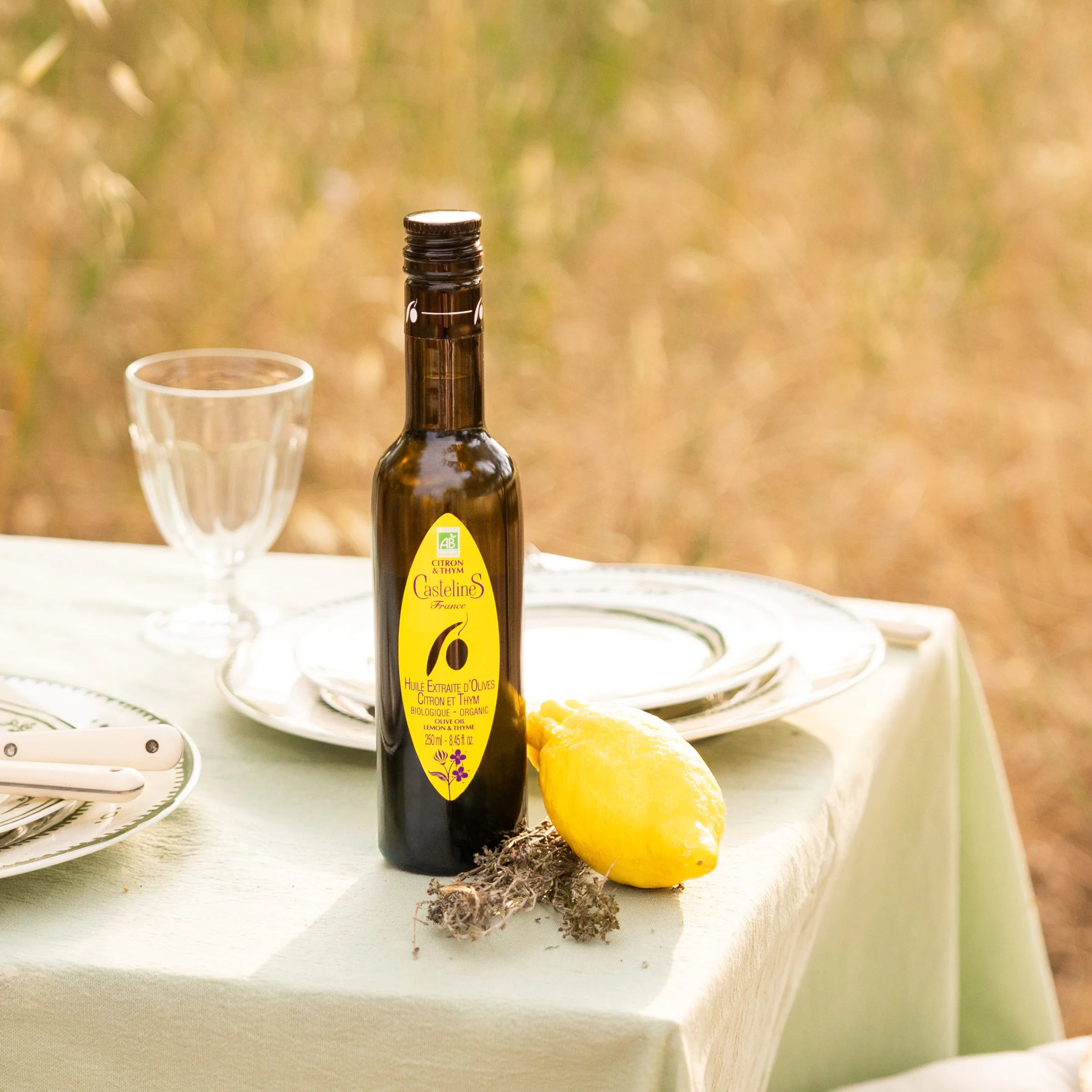 Close-up of Castelines lemon and thyme extra virgin olive oil bottle with fresh lemon and dried thyme on sage green tablecloth, Provence