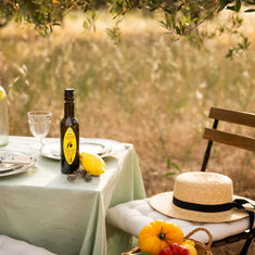 Castelines lemon and thyme olive oil bottle on outdoor Provençal table with sage green tablecloth, lemon, crystal glass and colourful tomatoes