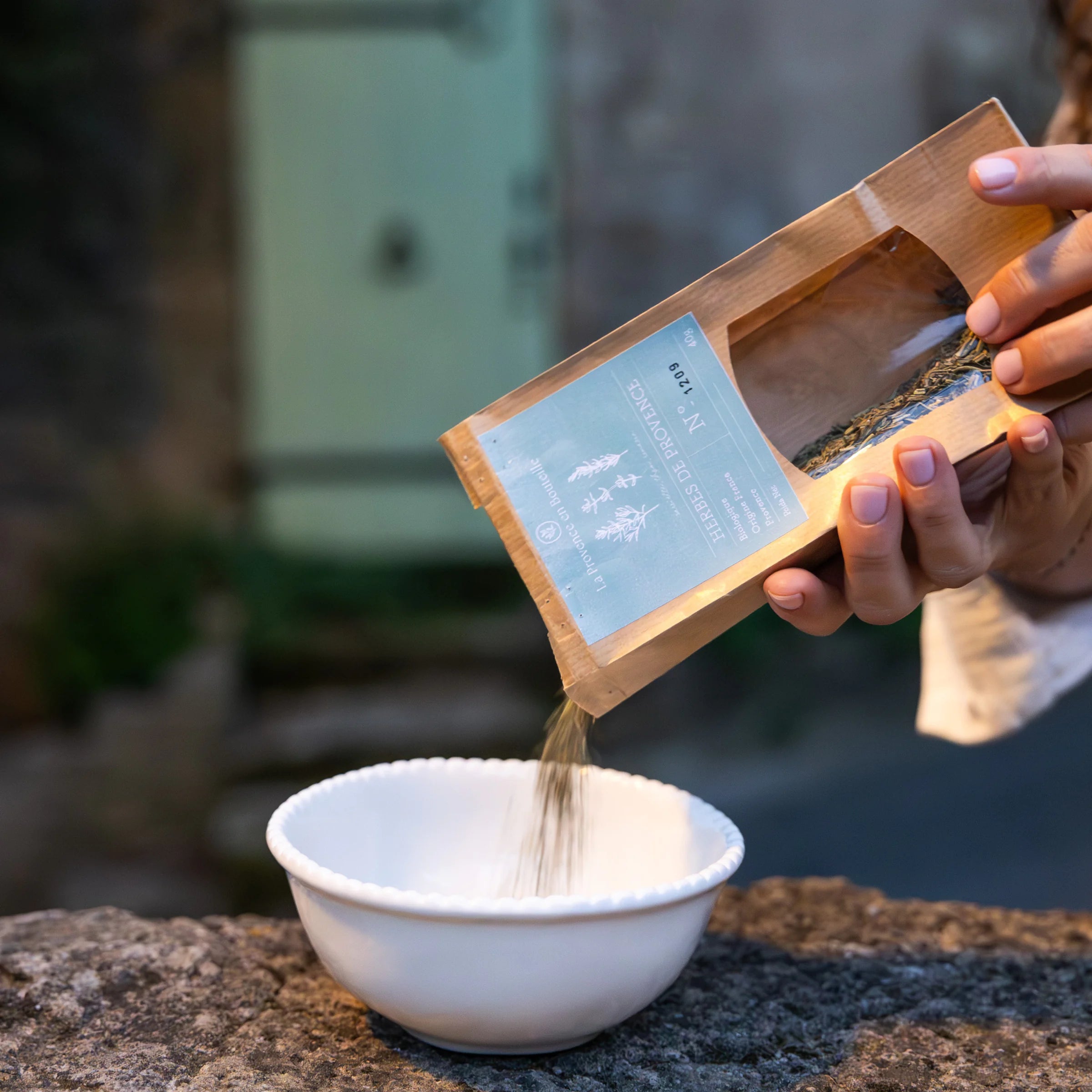 Hands pouring organic Provence herbs from La Provence en Bouteille bag into a white bowl, outdoors
