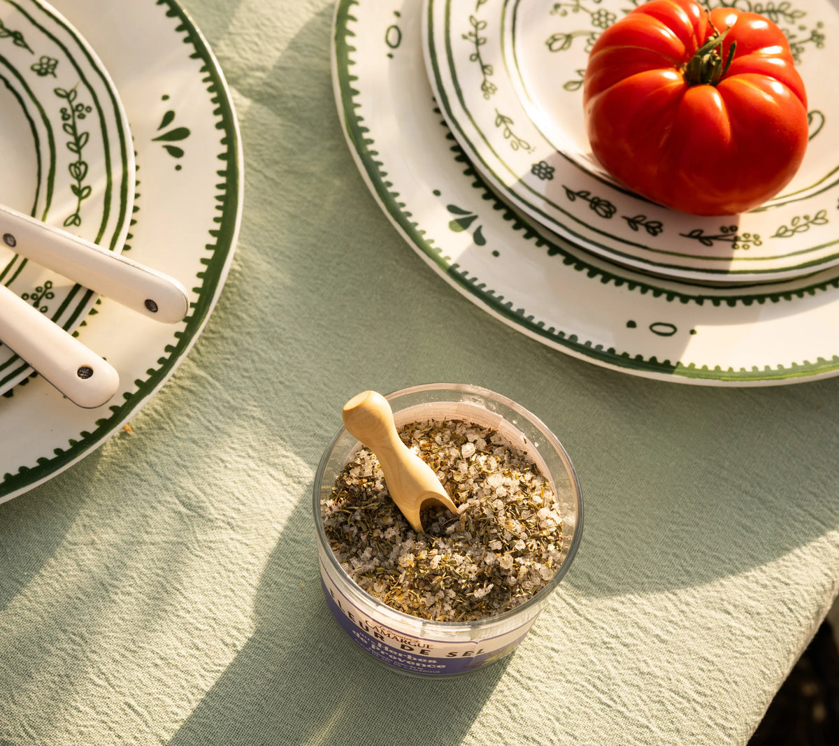Open Fleur de Sel with Provence herbs and wooden spoon on outdoor sage green Provençal table with green plates and fresh tomato, golden light

