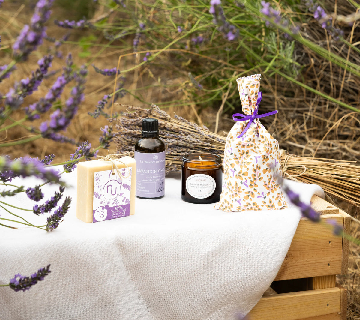 Four lavender products aligned on a linen wooden crate in a lavender field: lavender soap, Lavandin Grosso essential oil, lit lavender candle and floral lavender pouch

