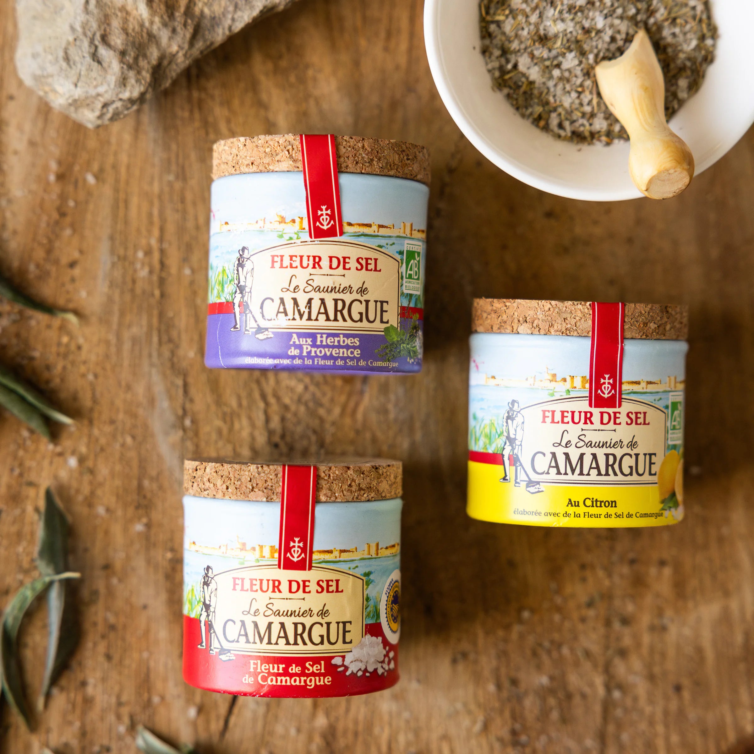 Hands stacking three Le Saunier de Camargue Fleur de Sel jars on stone, nature, lemon and Provence herbs trio
